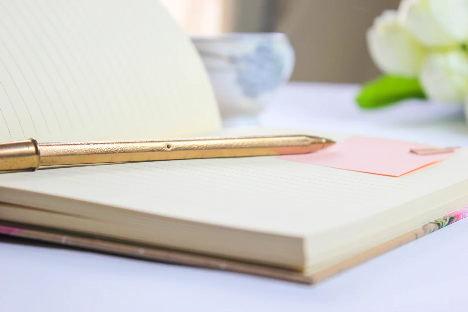 An open notebook with a gold pen and pink note alongside soft white flowers.
