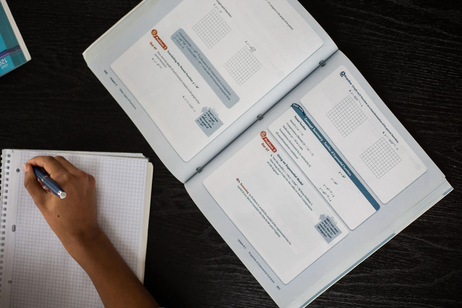 Overhead view of a person writing in a notebook beside open worksheet pages on a dark desk