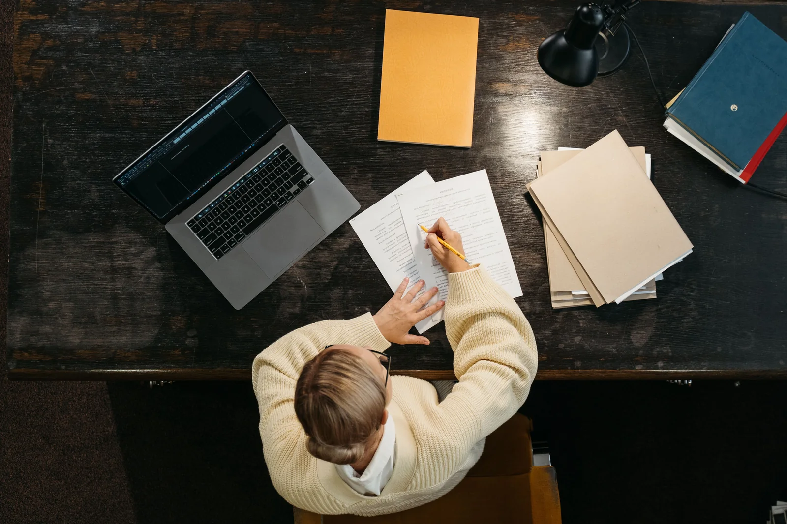 High-angle view of a writer reviewing and editing printed draft pages with a pencil beside an open laptop and folders on a wooden desk.