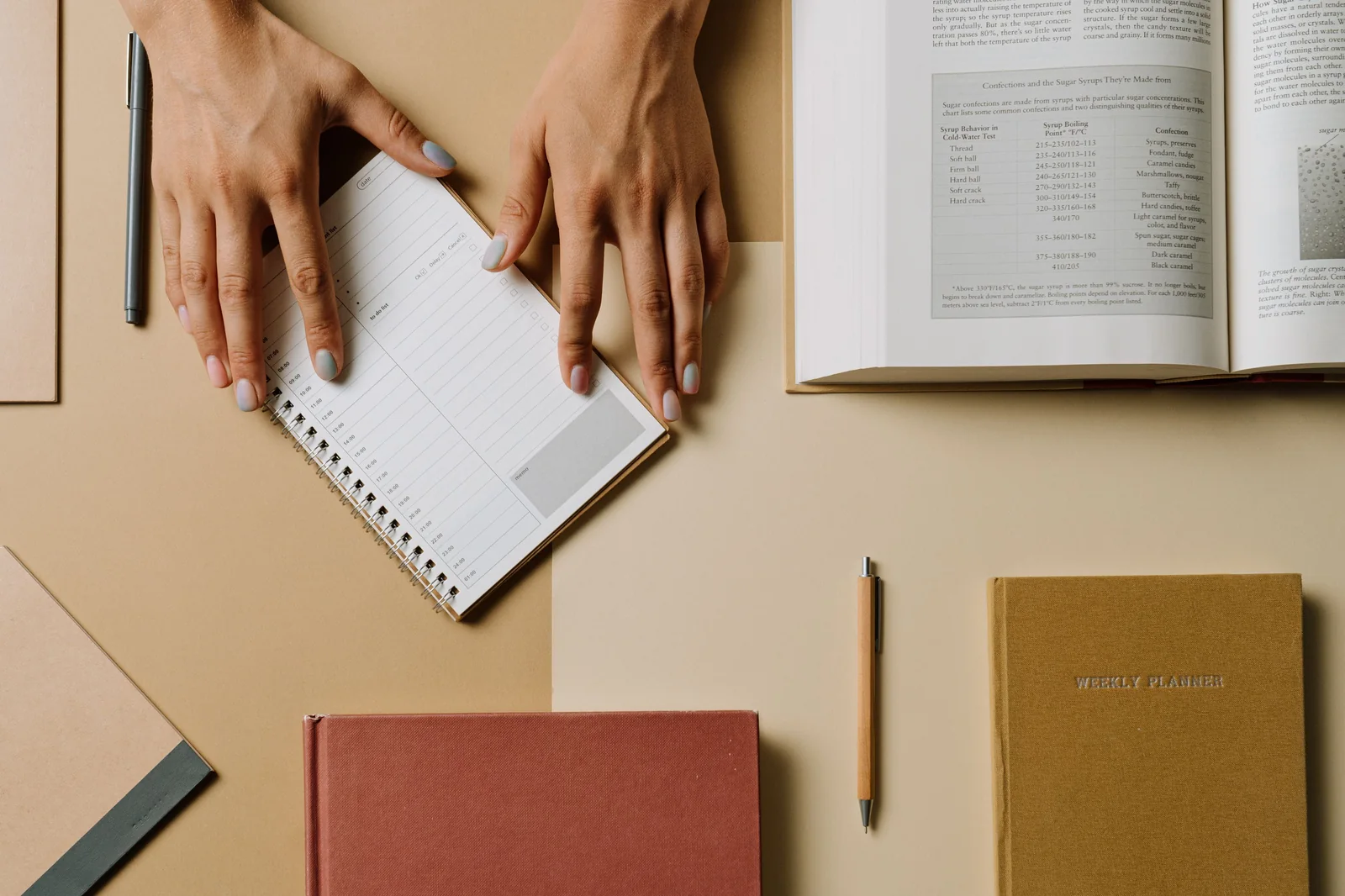 Top-down view of a writing desk with an open reference book, notebooks, pen, and hands organizing notes