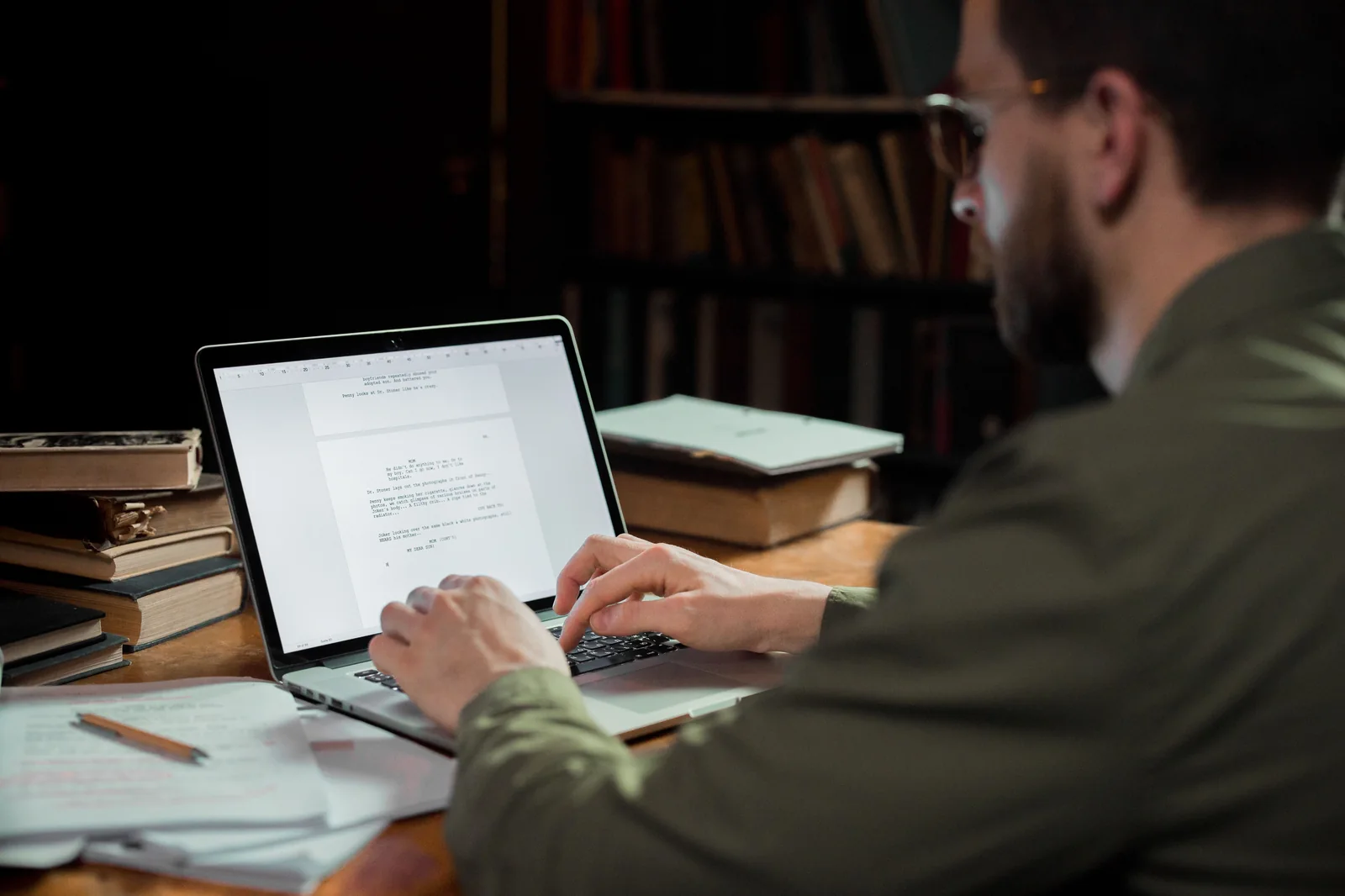 Over-the-shoulder view of an author typing on a laptop at a wooden desk with books and papers.