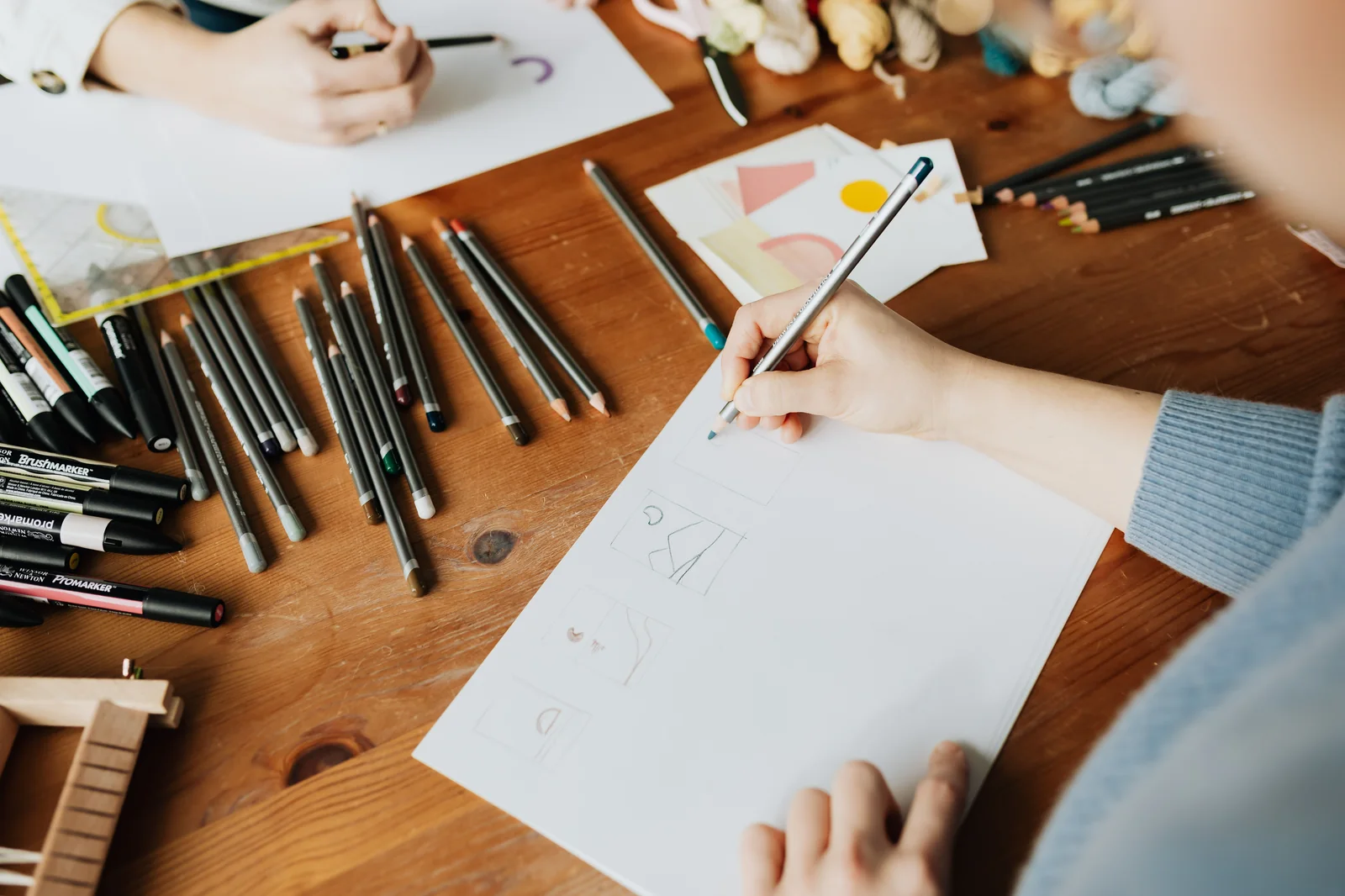 Overhead view of an illustrator's hands sketching storyboard panels on paper with colored pencils and markers on a wooden desk