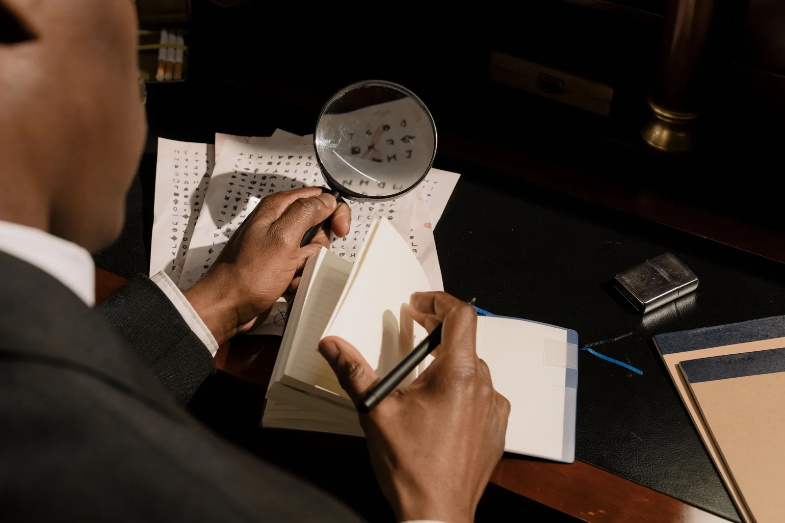A person reviewing cipher notes with a magnifying glass beside an open notebook at a dark desk.