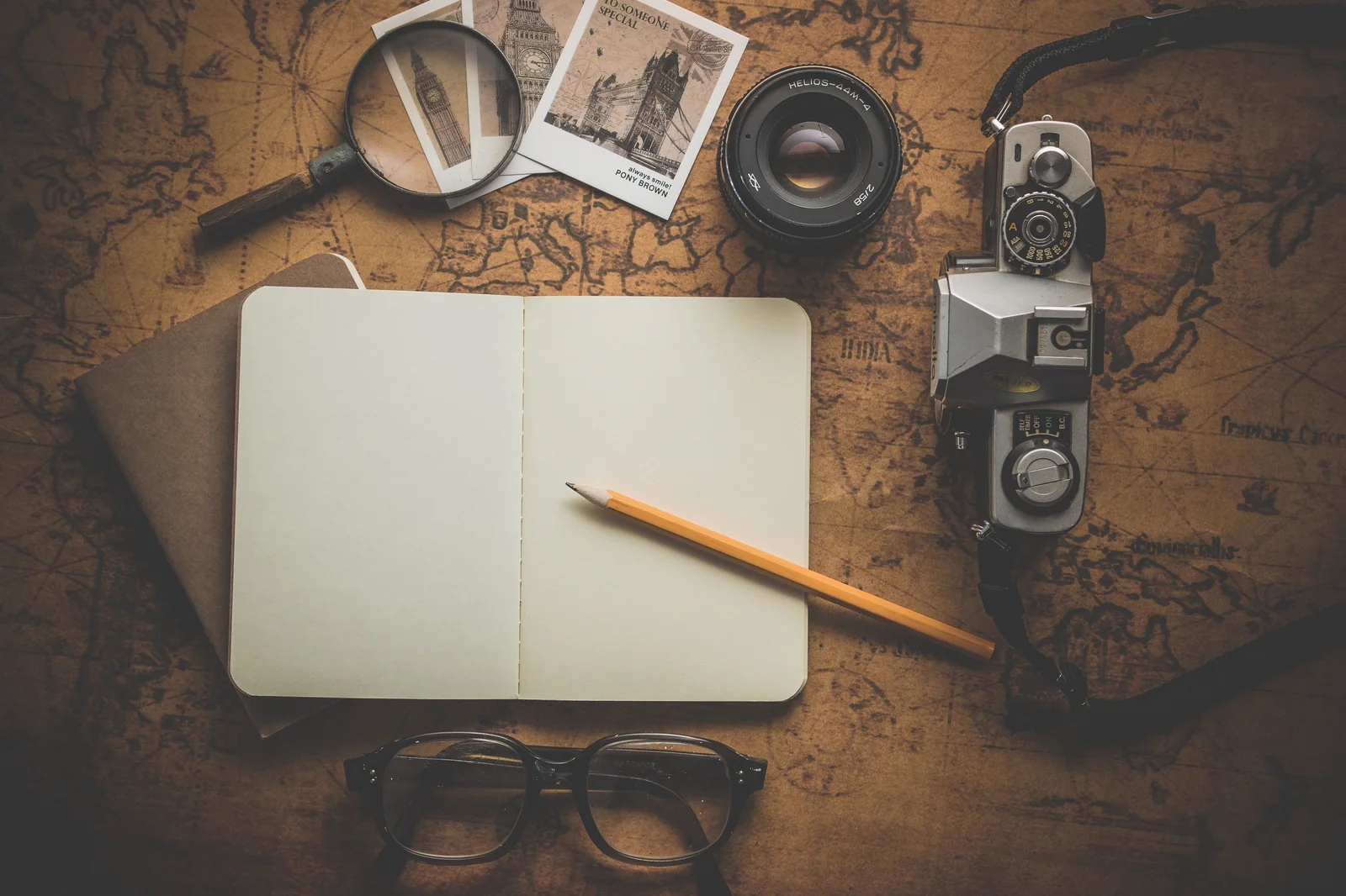 Flat lay of an open blank notebook, pencil, eyeglasses, and vintage camera over an antique world map