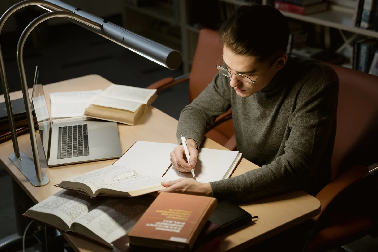 An academic taking notes at a desk surrounded by open reference books and a laptop under a lamp