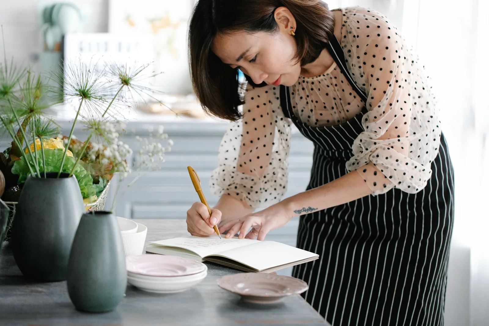 A woman wearing a striped apron writing in a notebook on a bright kitchen table