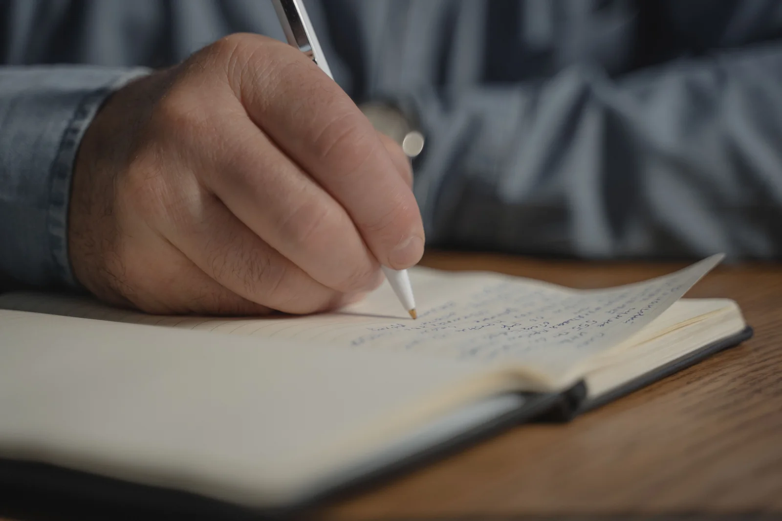 Close-up of a writer's hand drafting with a pen in an open notebook on a wooden desk