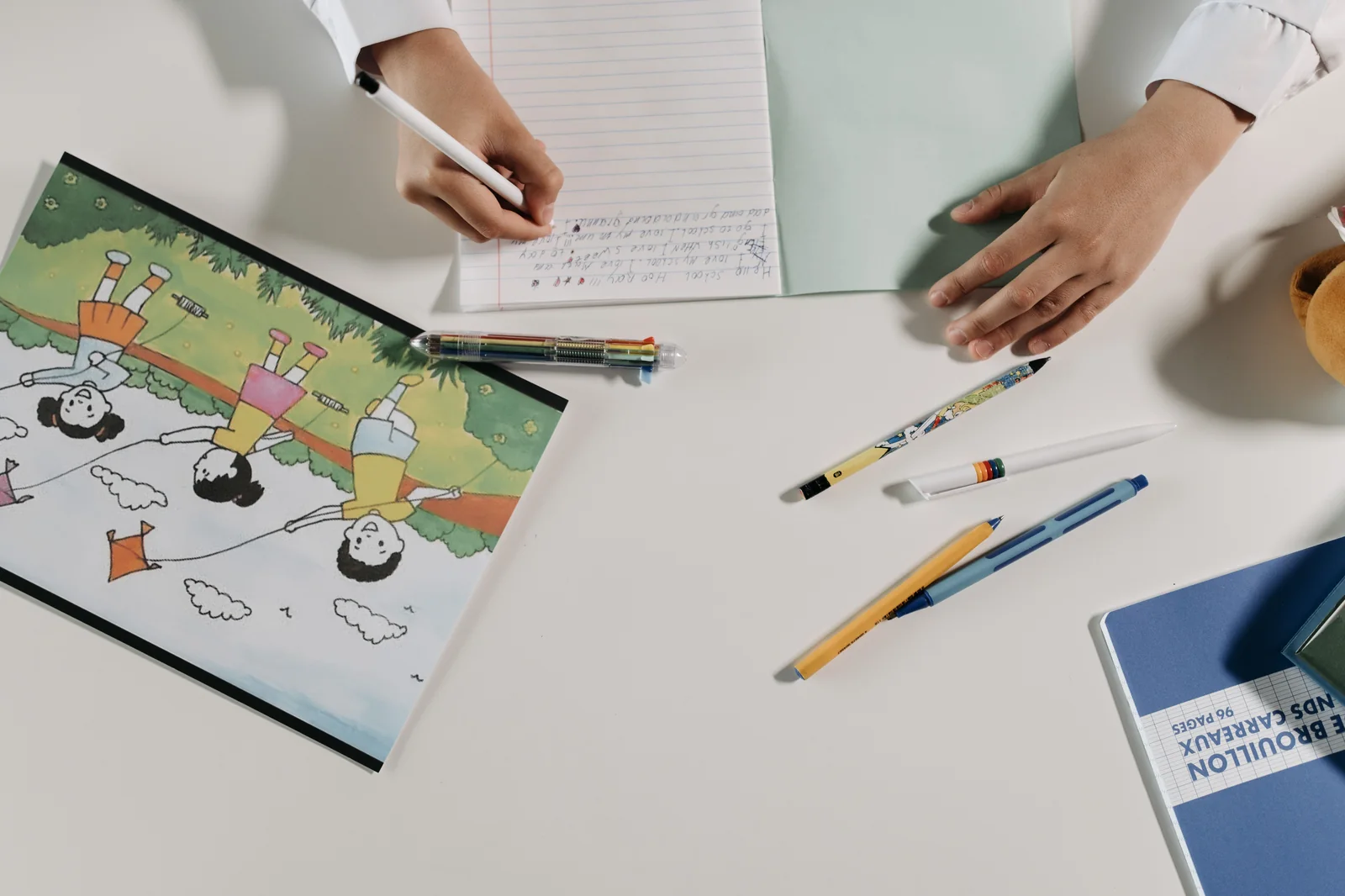 Overhead view of hands writing text in a notebook beside a colorful children's book illustration on a tidy desk