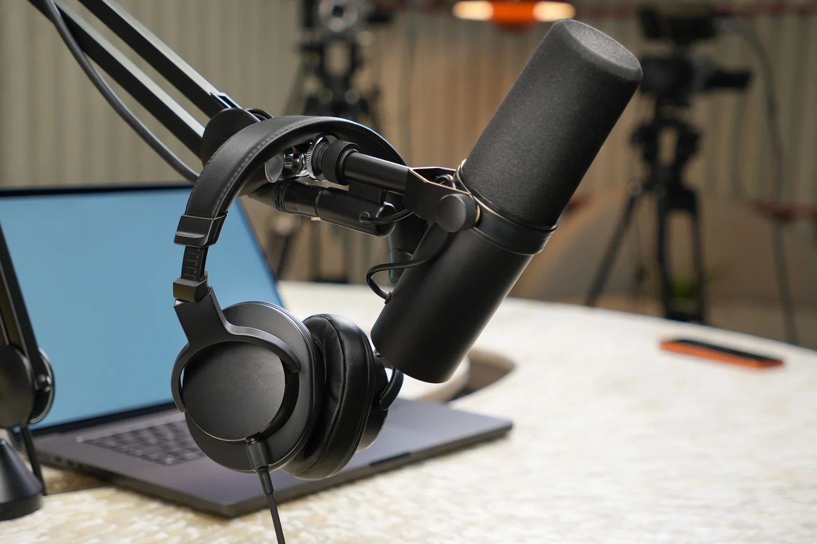 A black broadcast microphone and over-ear headphones on an adjustable boom arm beside a laptop on a desk.