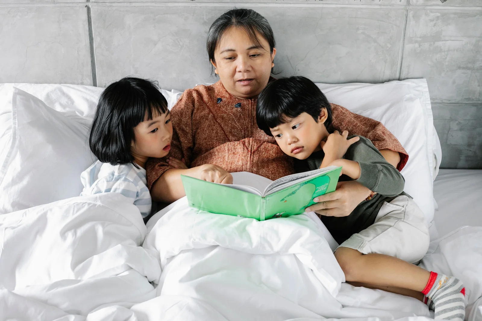 Mother and two young children reading an open storybook together in bed