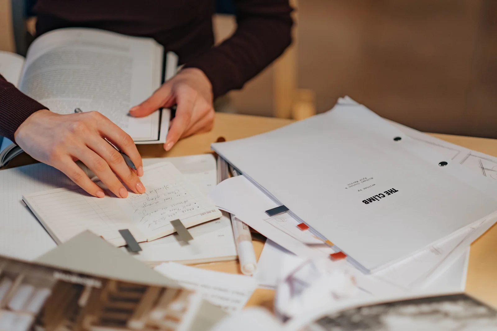 Hands taking notes in a notebook next to an open reference book and research papers on a desk