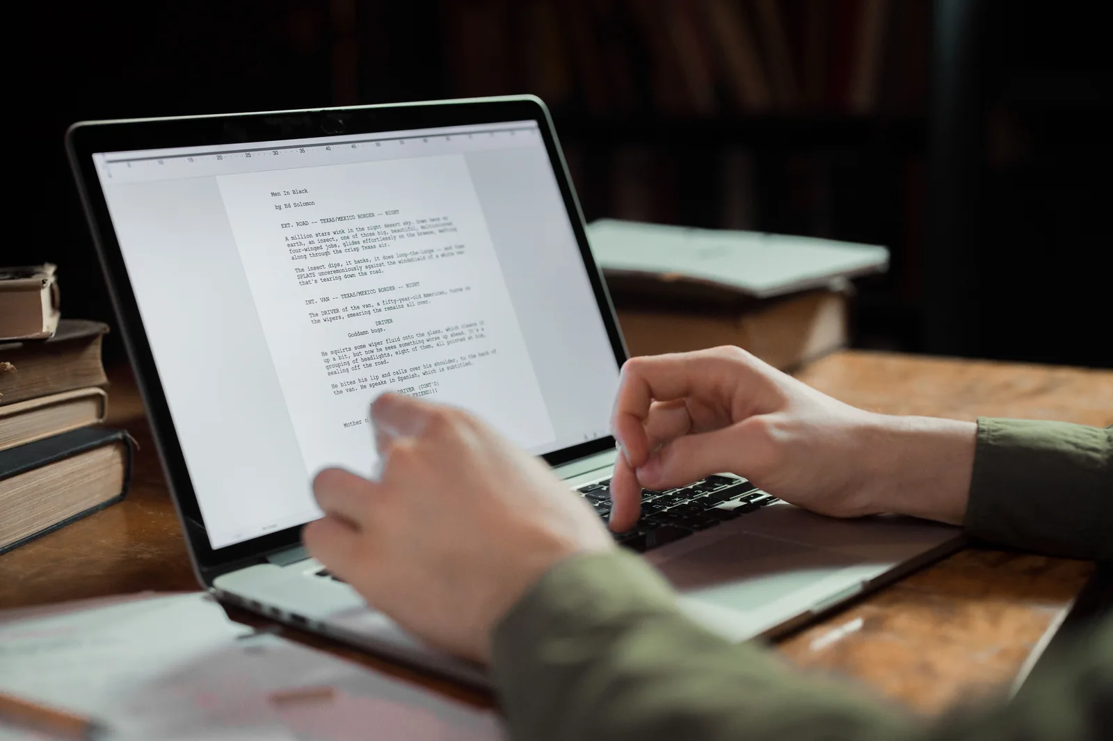 Over-the-shoulder view of a writer typing on a laptop at a wooden desk with books and papers