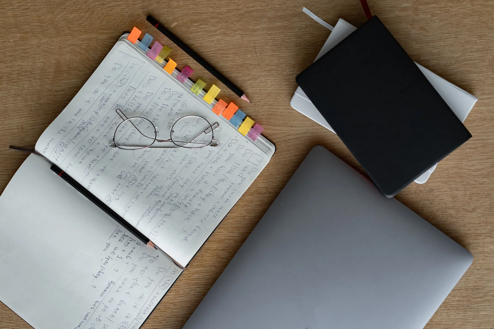 Overhead view of a wooden desk with an open notebook, colorful index tabs, glasses, notebooks, and a laptop.